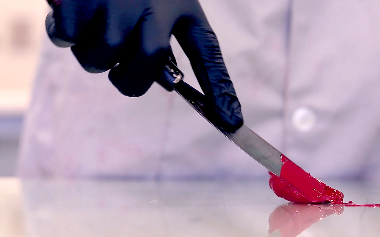 A person wearing a lab coat is using a spatula to mix red ink to print an engraving
