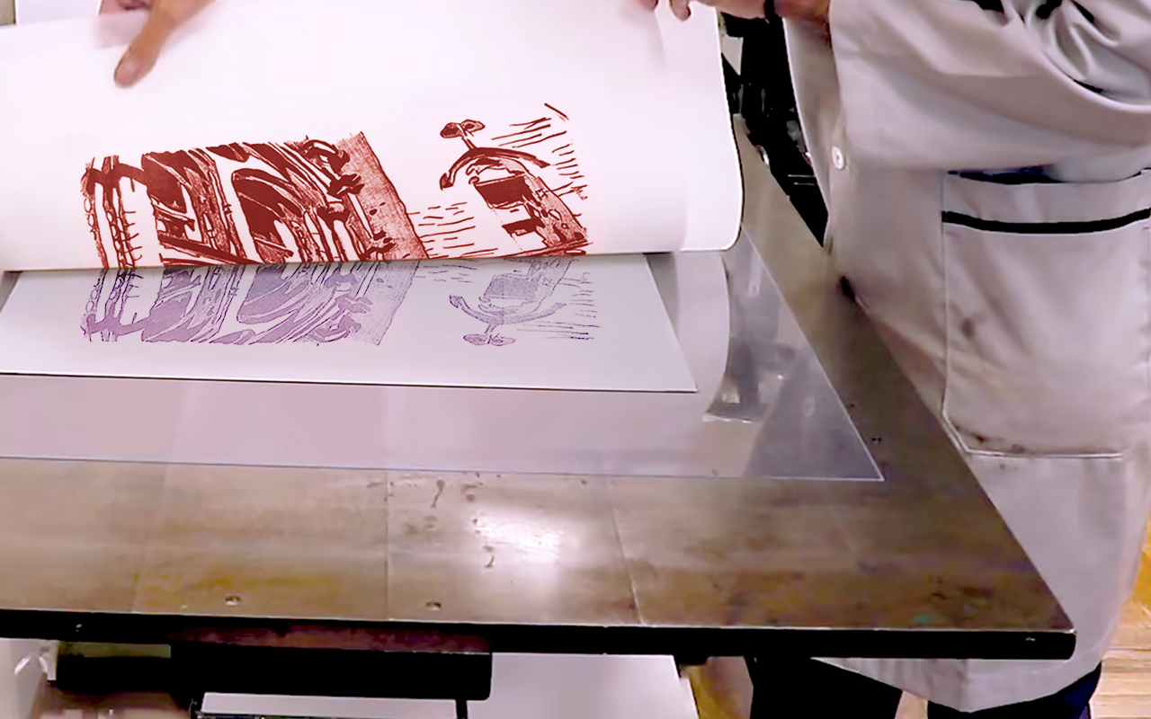 A man in an art lab coat carefully prints paper on an etching pres, focused on his task in a bright, organized workspace.